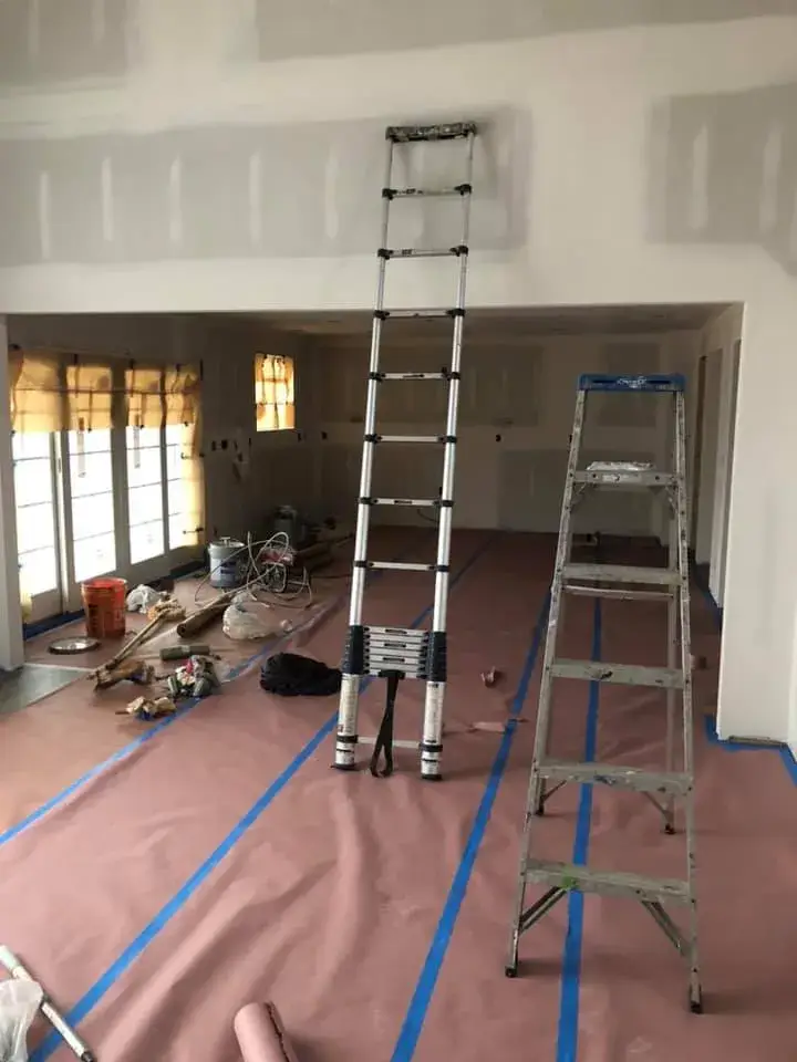 An extended telescoping ladder and a step ladder stand in a room under renovation with floors covered in protective paper. - My Personal Painter - My Personal Painter In progress: new-construction home interior painted