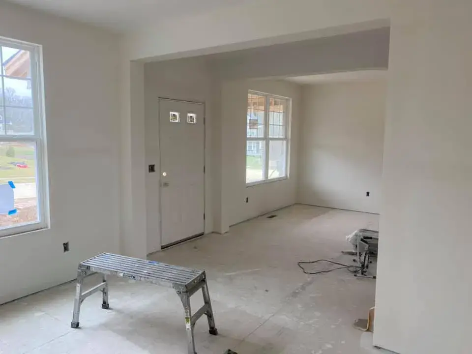 An unfinished room with white walls, windows, and a front door, featuring a metal step ladder on a concrete floor. - My Personal Painter - My Personal Painter In progress: new-construction interior walls painted