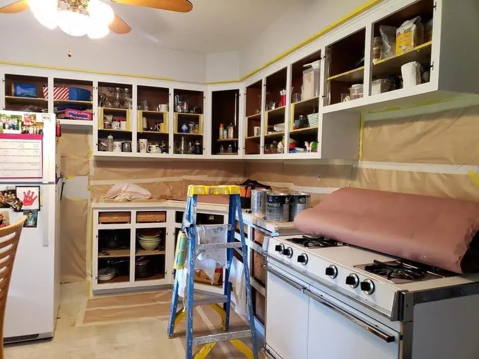 Kitchen cabinets with doors removed, masked with tape and paper for painting, featuring a step ladder in the center. - My Personal Painter - My Personal Painter In progress: oak kitchen: white uppers, dark gray lowers