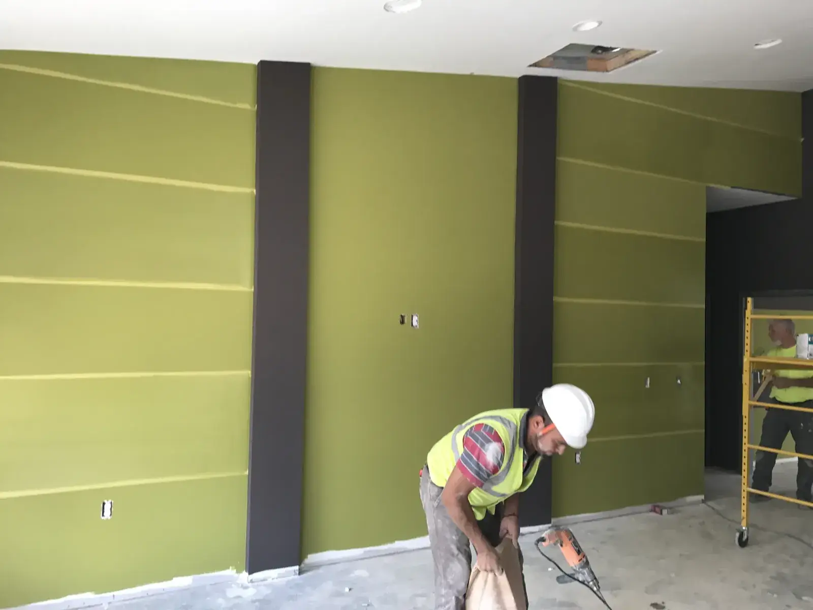 A construction worker uses a tool in front of a lime green wall with dark brown vertical panels. - My Personal Painter - My Personal Painter In progress: olive and charcoal striped commercial walls