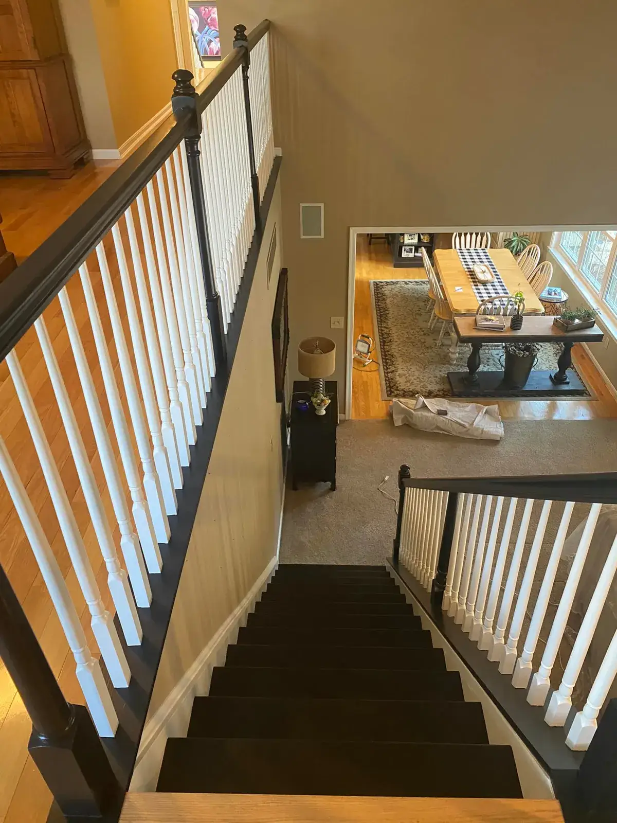 A view looking down a carpeted staircase toward a dining room with a long table and chairs. - My Personal Painter - My Personal Painter After: staircase: oak railing painted black with white spindles and black treads