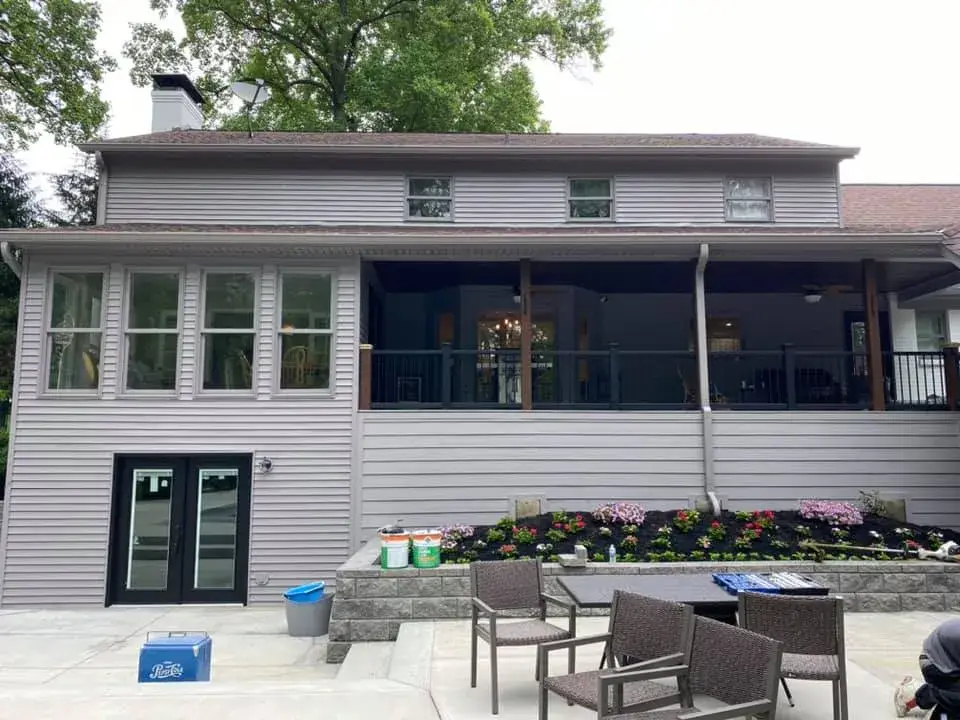The back of a two-story gray house with a covered deck, lower patio, and garden beds. - My Personal Painter - My Personal Painter After: two-story brick and siding house painted, red shutters to black