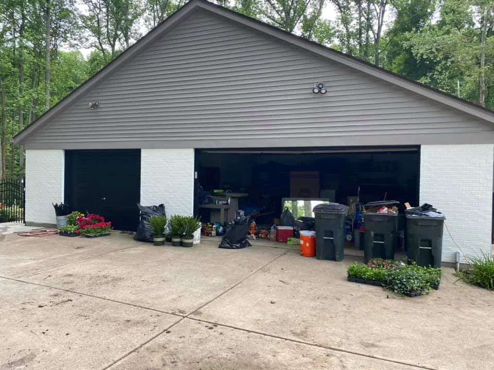 A two-car garage with one open bay, featuring potted flowers and trash bins on a concrete driveway. - My Personal Painter - My Personal Painter After: two-story brick and siding house painted, red shutters to black