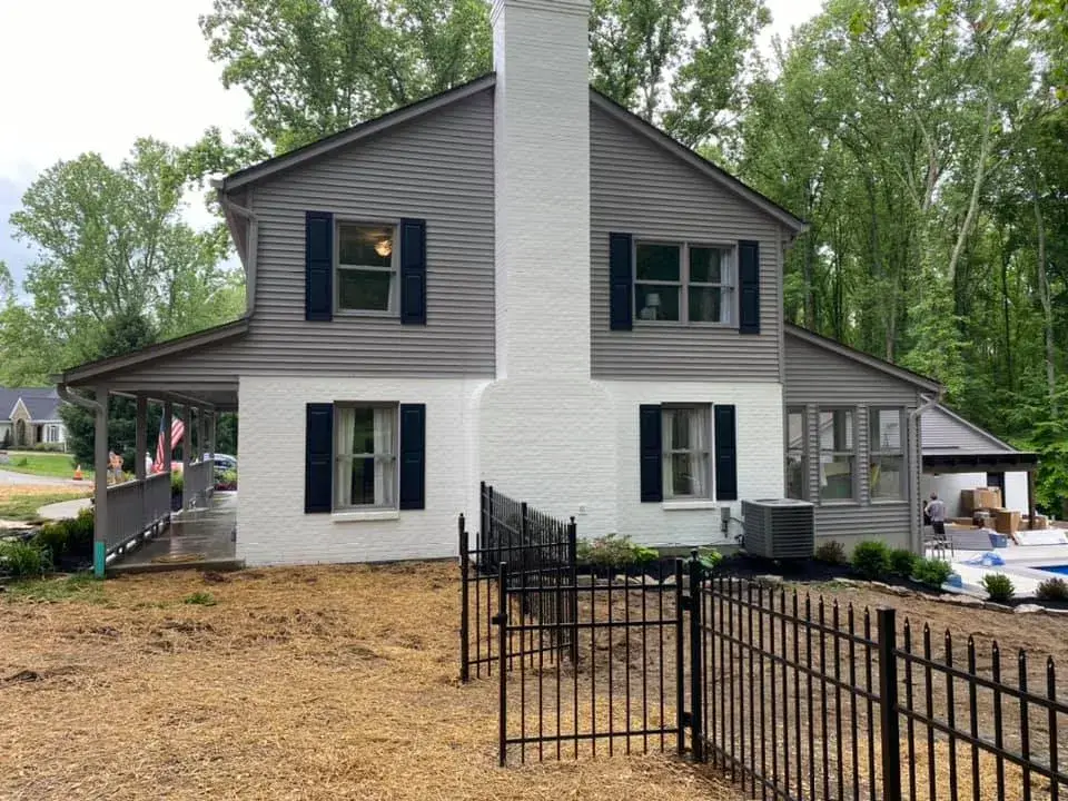 Two-story house with gray siding, white brick chimney, and black shutters, featuring a wraparound porch and fenced yard. - My Personal Painter - My Personal Painter After: two-story brick and siding house painted, red shutters to black