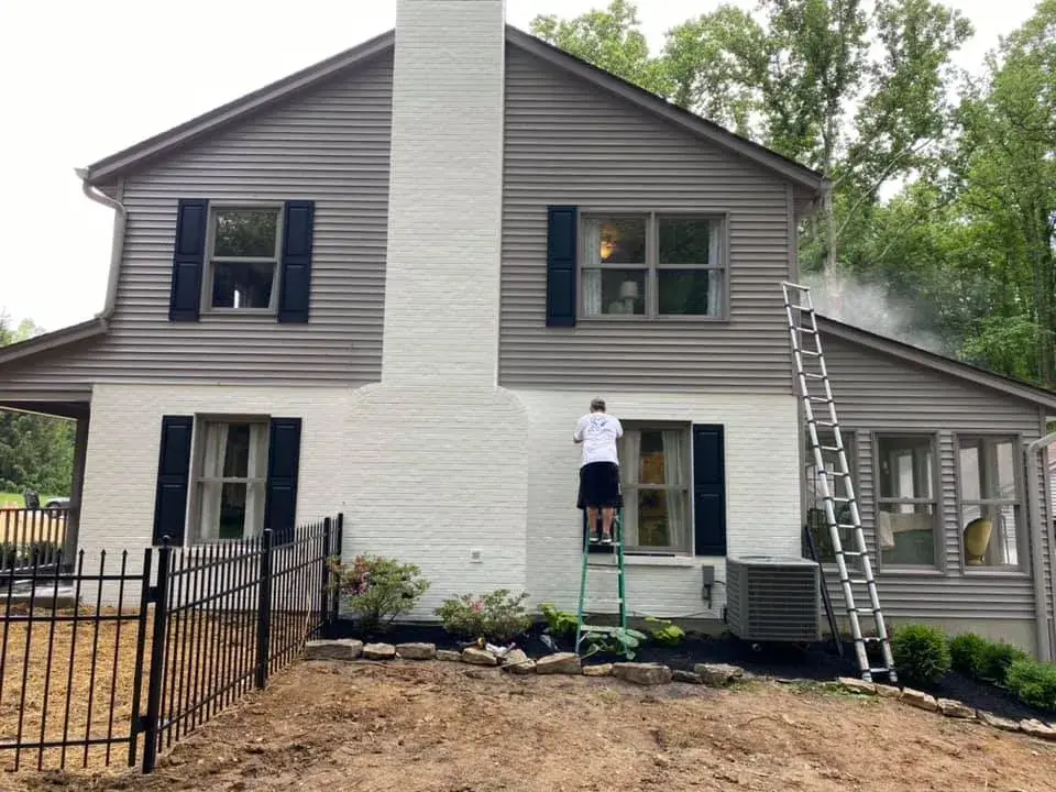 A person stands on a ladder to paint a white brick chimney on the side of a gray house. - My Personal Painter - My Personal Painter In progress: two-story brick and siding house painted, red shutters to black