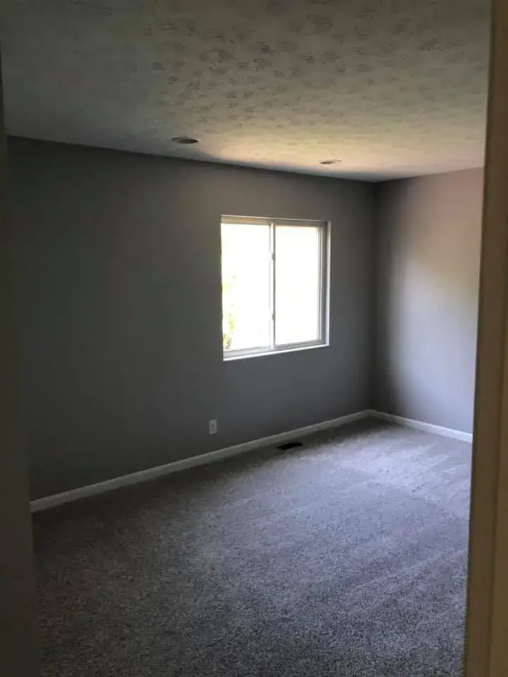 An empty bedroom with gray walls, light-colored carpeting, and a single window. - My Personal Painter - My Personal Painter After: two-story foyer and rooms painted gray, stair railing painted black