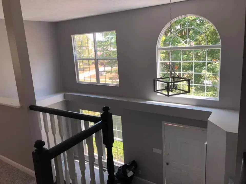 A view from a staircase landing looking down toward a front door with large arched and rectangular windows. - My Personal Painter - My Personal Painter After: two-story foyer and rooms painted gray, stair railing painted black