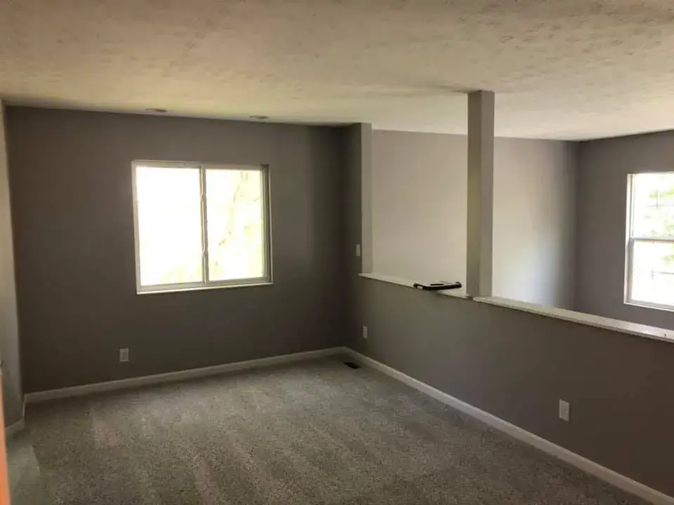 Empty room with gray walls, carpeted floor, two windows, and a half-wall partition. - My Personal Painter - My Personal Painter After: two-story foyer and rooms painted gray, stair railing painted black