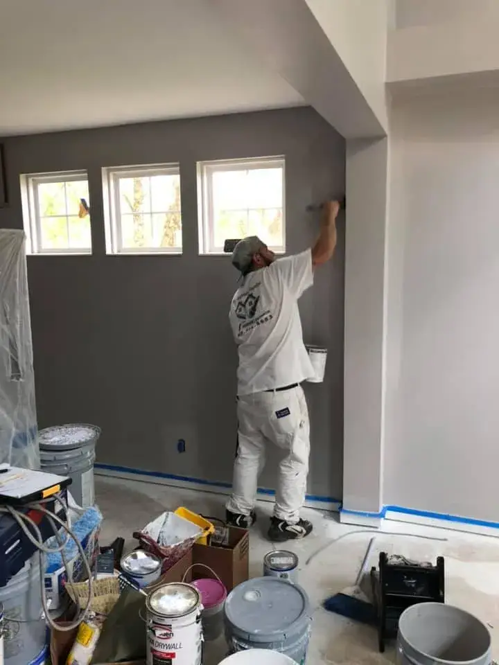 A painter wearing white work clothes paints a gray interior wall near windows. - My Personal Painter - My Personal Painter In progress: two-story foyer and rooms painted gray, stair railing painted black