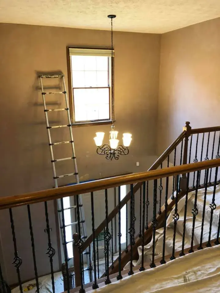 An extension ladder leans against a wall in a two-story foyer near a chandelier and a staircase. - My Personal Painter - My Personal Painter In progress: two-story foyer with iron balusters painted