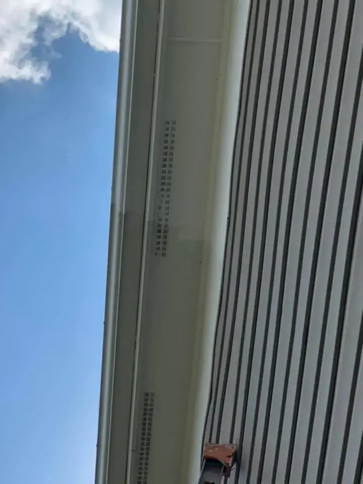 A low-angle view of a house eave with a white vented soffit against grey siding and a blue sky. - My Personal Painter - My Personal Painter In progress: two-story tan siding home exterior trim