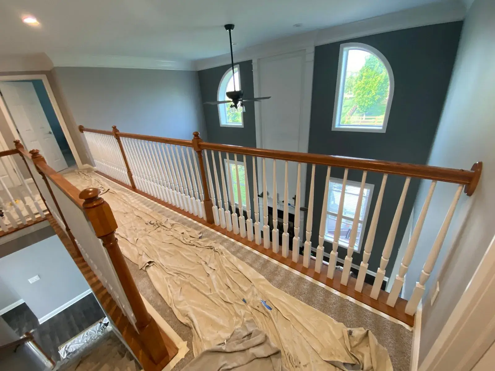 A wooden staircase railing overlooks a two-story living area with dark grey walls and large arched windows. - My Personal Painter - My Personal Painter Before: upstairs railing painted: black handrail, white spindles