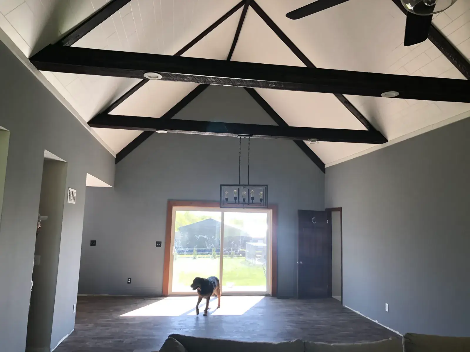 A dog stands in a vaulted room with dark wood beams and a large glass door leading to a backyard. - My Personal Painter - My Personal Painter After: vaulted great room: brick chimney painted, then stained wood ceiling to white planks and black beams