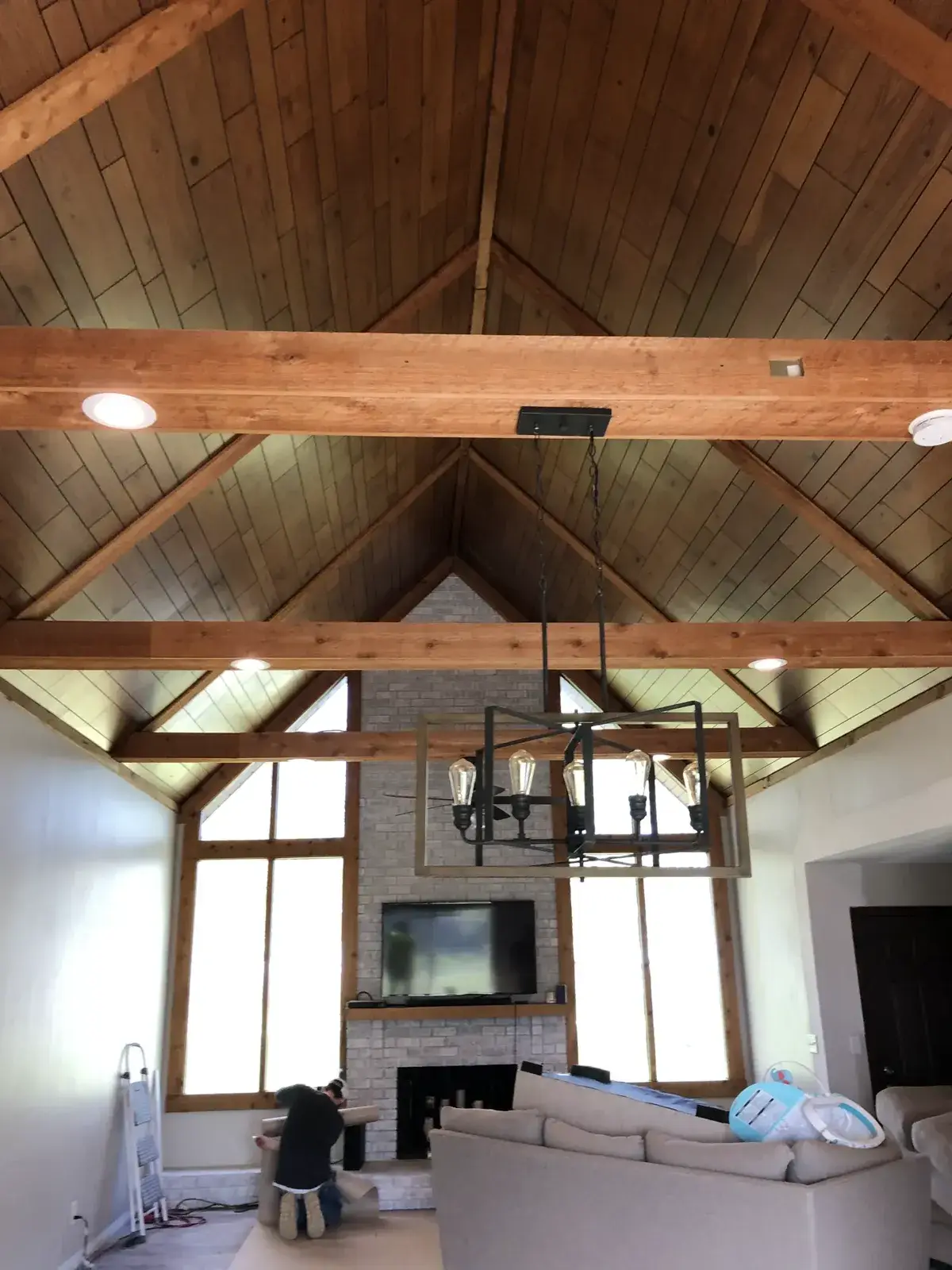 A room with a high, wood-paneled vaulted ceiling, exposed beams, a rectangular chandelier, and a stone fireplace. - My Personal Painter - My Personal Painter Before: vaulted great room: brick chimney painted, then stained wood ceiling to white planks and black beams