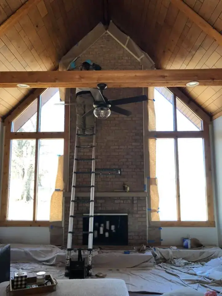 A person on a tall ladder masks a brick fireplace with paper in a room with a vaulted wood ceiling. - My Personal Painter - My Personal Painter In progress: vaulted great room: brick chimney painted, then stained wood ceiling to white planks and black beams