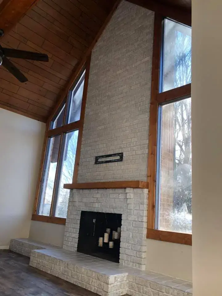 A tall, light-colored brick fireplace with a wood mantle, flanked by large windows reaching up to a vaulted wood ceiling. - My Personal Painter - My Personal Painter In progress: vaulted great room: brick chimney painted, then stained wood ceiling to white planks and black beams