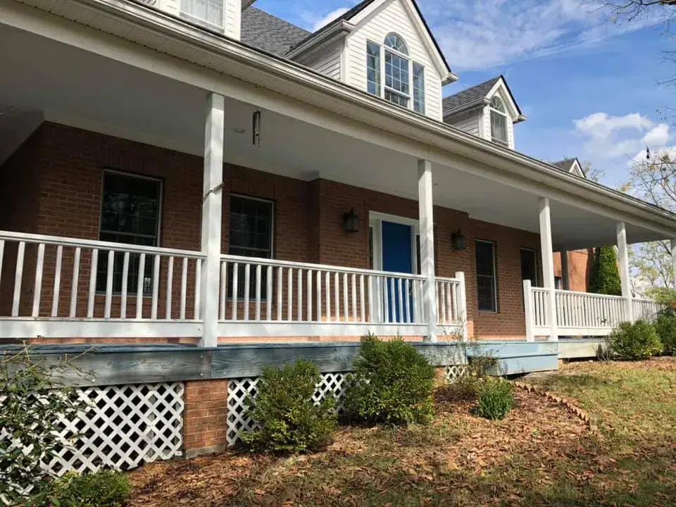 A brick house with a wide covered front porch, a blue door, and white wooden railings. - My Personal Painter - My Personal Painter After: wraparound porch on brick Cape Cod: railings white, floor painted blue-gray